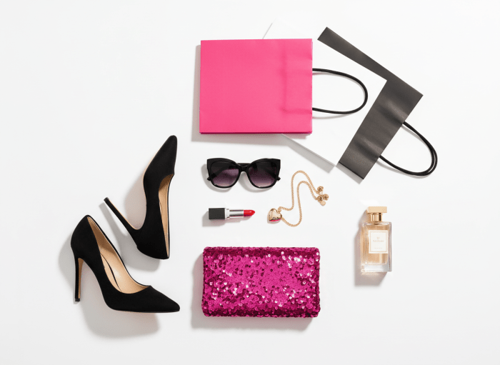 A beautiful fashion flat lay on a pure white background. Arranged artfully: a red lipstick, a delicate gold chain necklace with a heart pendant, stylish black oversized sunglasses, a hot pink sequin sparkle clutch purse, two pairs of black stiletto heels, a glass bottle of perfume with gold cap. Also included: a plain hot pink shopping bag, a white shopping bag, and a black shopping bag. All items are neatly spread out in an editorial flat lay style with soft shadows, clean and luxurious feel.