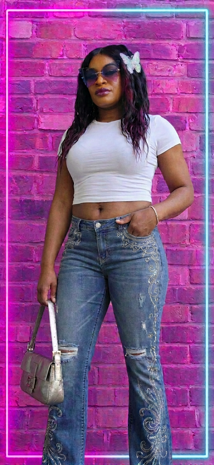 Woman wearing sunglasses, a white crop top, embellished jeans, and holding a handbag before a pink brick wall