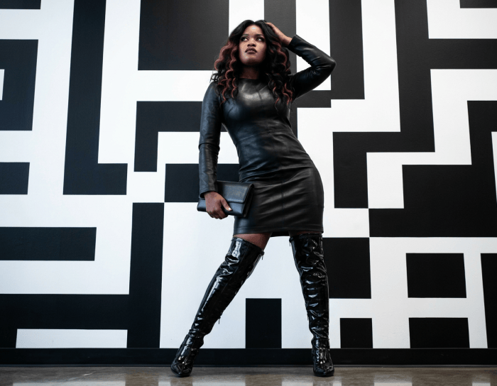 Woman in a black leather dress and thigh-high boots posing against a geometric black-and-white wall
